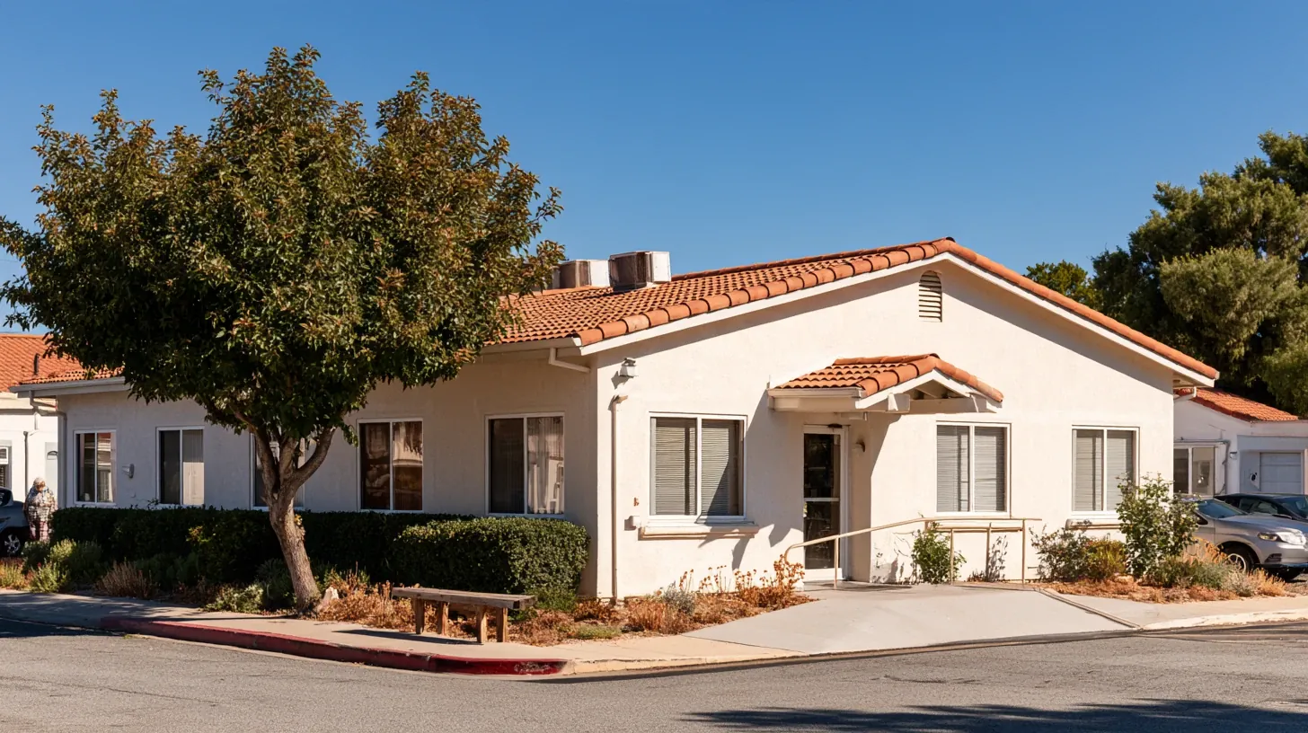 Exterior view of a single-story assisted living building on a bright sunny day, featuring light stucco walls, red tile roofing, and an accessible wheelchair ramp leading to the front entrance. A wooden bench sits beneath a green tree in front of manicured hedges, with a clear blue sky in the background.